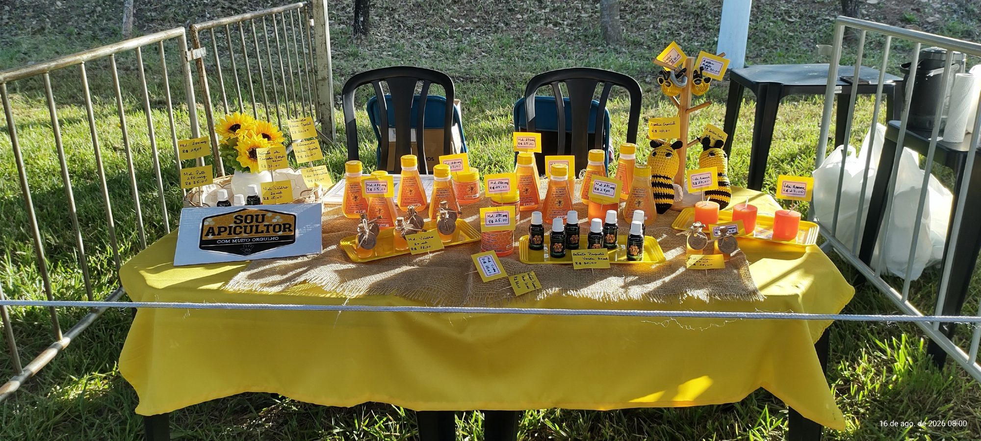 Vista geral da mesa da Colmeia montada na feira, com toalha amarela e produtos expostos.